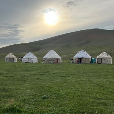 Six yourtes blanches sur un pré verdoyant, avec un soleil couchant derrière une colline.