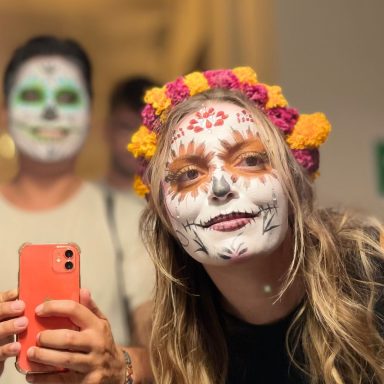 Femme souriante avec maquillage coloré et couronne de fleurs, un homme derrière elle.