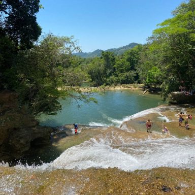 Cascade d'eau claire avec des personnes se baignant dans une piscine naturelle entourée de verdure.