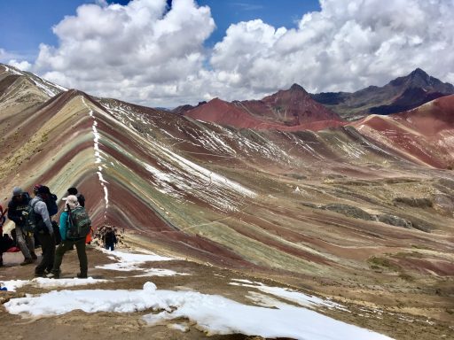 Randonneurs sur un sommet avec des montagnes colorées sous un ciel nuageux.