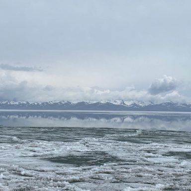 Vue d'un lac calme avec des montagnes enneigées à l'horizon sous un ciel nuageux.