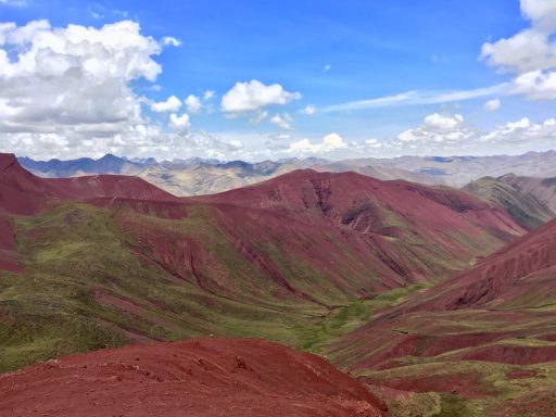 Vue panoramique de montagnes rougeâtres sous un ciel nuageux avec des vallées verdoyantes.