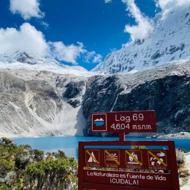 Vue d'un lac turquoise en montagne, avec un panneau d'information et des sommets enneigés.