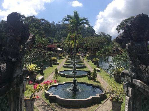 Jardins avec fontaines et palmiers, entourés de verdure sous un ciel bleu.